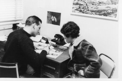 A man sits at an office desk and points to a page in a book. A woman sits beside him and looks to where he is pointing.