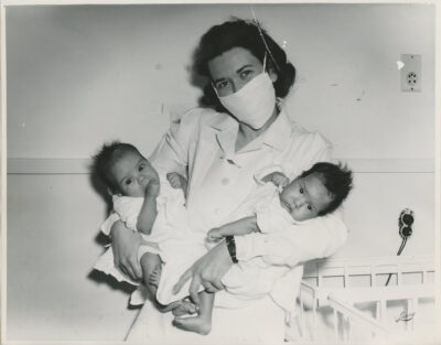 A nurse wearing a face mask holds two babies.