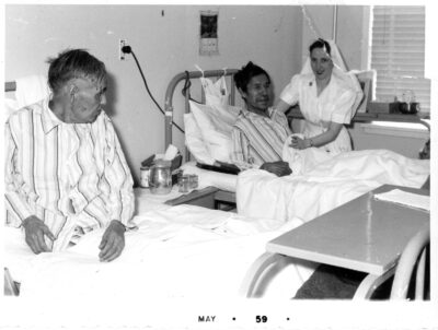 Two men wearing striped pyjamas in hospital beds. A nurse tucks one of the men into bed.