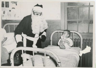 A man dressed as santa claus stands next to a toddler in a hospital bed. Another child can be seen in a bed at the edge of the frame.
