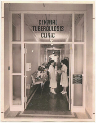 The entrance of the Central Tuberculosis Clinic. Two nurses stand in the hallway and a group of people sit on chairs against the wall.