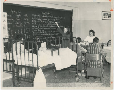 A classroom with some students in hospital beds and some in chairs. One student writes on a chalkboard for a "silent reading test" while the teacher watches from the front of the room. A framed photograph on the wall above the teacher can be found in the Grey Nuns Archives, no. 22-25.7