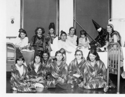 A group of women in costumes. Some women are sitting in hospital bends, and some are sitting in front on the ground.