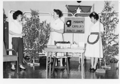 A woman speaks to another woman by a table with beauty products on it. Another woman in a maid's uniform, holding a mop stands by the table. A sign behind the table reads, "Madame Camelia's Beauty Shop" and Christmas trees and decorations are placed around the scene. Caption on reverse reads, "Adult Party"