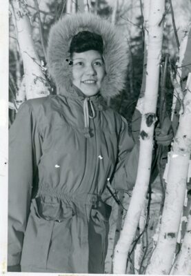 A woman in a fur-trimmed parka outside amongst trees. Reverse shows stamp that reads, "Aurora Photo Studio, 205 Fisher Avenue, The Pas Manitoba"