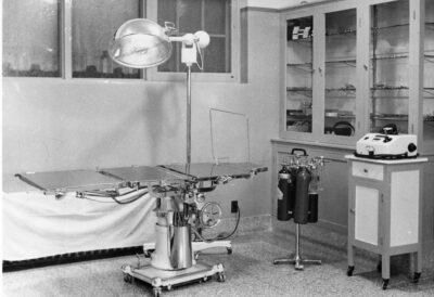 A view of a procedure room with a metal operating table, large lamp, and a stand containing metal tanks. From album page MBLung-11-15-001