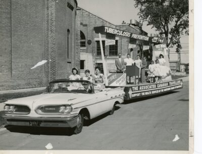 Three women sit on the back seat of a convertible car, which pulls a parade float. Adults and children stand on the float which has a sign that reads, "The Associated Canadian Travellers Support the Health Services of the Sanatorium Board of Manitoba"