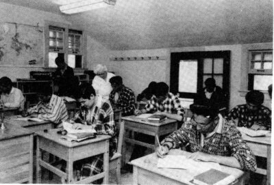 Men in patterned pyjamas sit at individual desks and write. A woman stands next to one man and looks at his work.