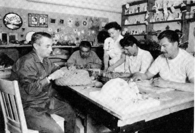 Four men sit around a table and work on handicrafts. A woman stands next to them and assists.