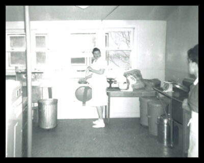 A nurse appears to be baking in a kitchen. She looks at another nurse at the edge of the frame. Scales, large sacs, and a stand mixer sit on a counter.