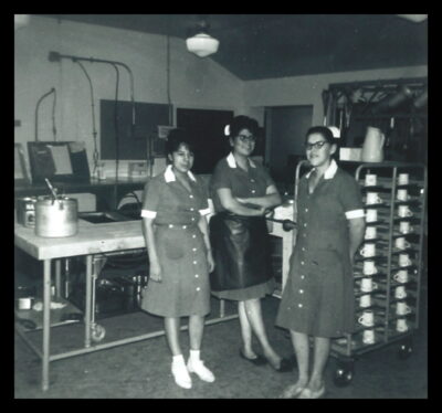 Three women stand in a dish-washing room, next to a rack of mugs.