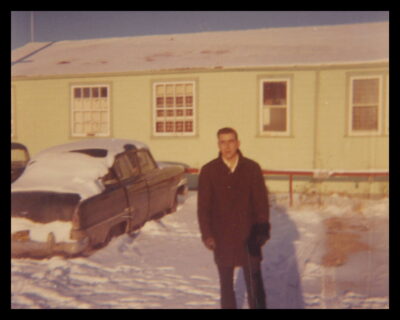 A man stands in a snowy parking lot by a single-storey building. A car covered in snow is parked next to him.