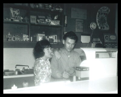 A man and woman stand behind the counter of a store. The woman rests her arm on the man's shoulder.