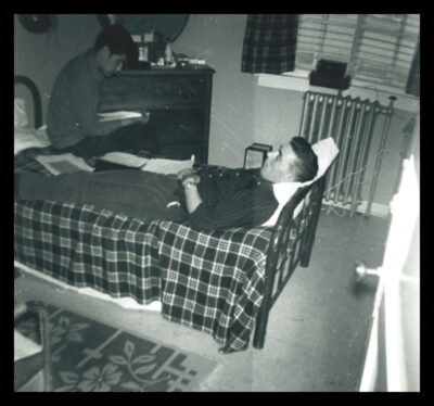 A man lies on a bed with a plaid blanket, and another man sits on the bed next to him, reading a book.