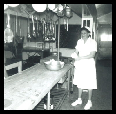 A nurse stands in a kitchen, next to a row of hanging pots and utensils.