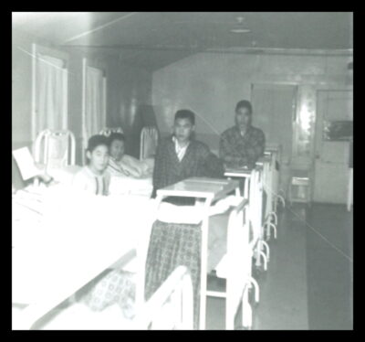 A row of hospital beds with young male patients. Two young men in plaid robes sit on the ends of two of the beds.
