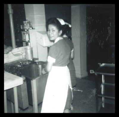 A woman stands at a sink and turns towards the camera. Another woman can be partially seen next to her.