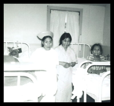 A woman in a nurse's cap and another woman stand between two patient beds. A woman lies in the bed on the left and another woman sits in bed on the right.