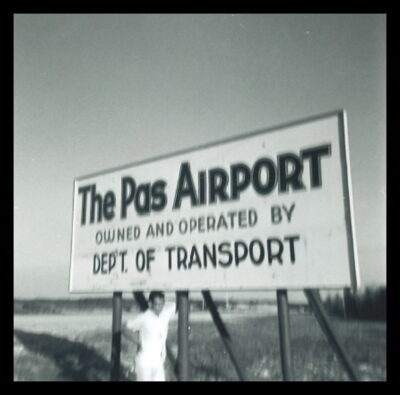 A man stands under a sign that reads, "The Pas Airport Owned and Operated by Dept. of Transport"