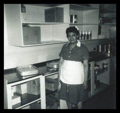 A woman stands next to a counter with trays, and shelves holding boxes and large cans