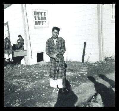 A man in a plaid robe stands outside near a building. Two people can be seen sitting on the landing of the building.