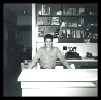 A man stands behind a counter. The shelves behind him are filled (contents unclear) and there are coffee pots on a hot plate on the counter behind him.