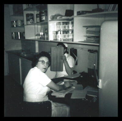 Two women sit at a table under shelves filled with salt boxes and large cans. One woman looks at the camera with a surprised expression, and the other woman shields her face with a clipboard.