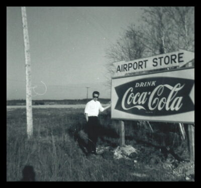 A man stands by a sign that reads, "Airport Store // Drink Coca Cola"