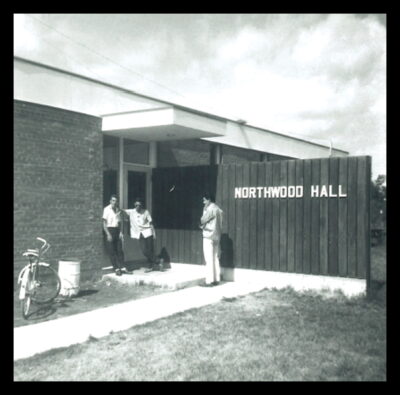 Three men stand at the entrance of a building. A sign reads, "Northwood Hall."