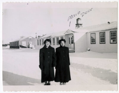 Two ladies in long black coats are standing in front of the Clearwater Lake Indian Hospital building in winter time.