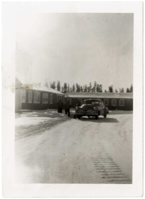 A taxi is parked outside the staff quarters of the Clearwater Lake Indian Hospital picking up two passengers. The driver is outside of the car.