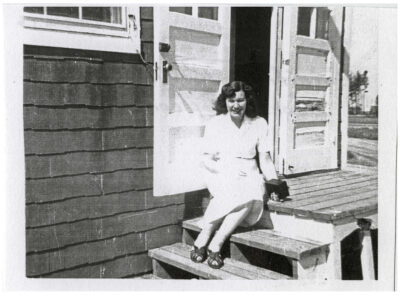 A woman is sitting on the doorstep of the "Girls Quarters" at the Clearwater Lake Sanitorium.  She is holding a camera in her hand.  The steps she is sitting on leads to two doors.