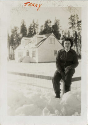 A woman is sitting on a fence in winter. A house is in the background along with some trees, snow on ground.