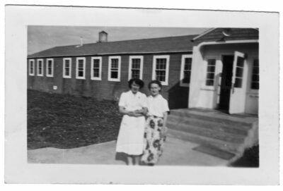 Two women link arms outside of a building with an open door. One woman wears a patterned skirt and the other woman wears a nurse's outfit.