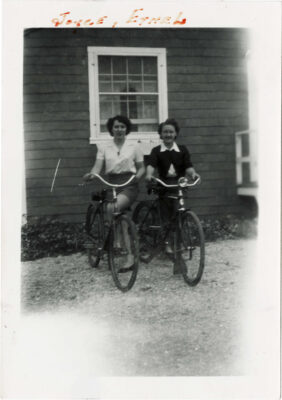 Two women stand with bicycles next to a window of a building. Shrubs line the bottom of the building.