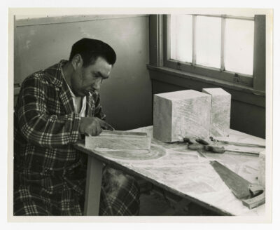 A man preparing a piece of soapstone for carving. He is handling a file or rasp on the smallest piece of soapstone in the image. There are three other pieces of soapstone visible. Two saws are visible on the work table. He has a cigarette in his mouth and is wearing pyjamas and a plaid patterned housecoat. The table he is working at is covered in soapstone dust. He is sitting beside a closed window.