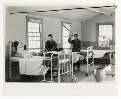 Three patients in a hospital ward. One man is standing, facing the camera and waving while smiling. Another is sitting on a bed and facing the camera but looking down at another man. The other man is seated on a chair facing away from the camera and towards the other two. All three are dressed in pyjamas and plaid patterned housecoats. There are three beds visible, white metal frames with thin mattresses and wheels, two pillows each and white sheets. The closest bed has a metal eating tray rolled over the bed and a pair of shoes are on top. There are a line of tables to the PL of the image with a water jug visible. There are three windows visible in the image with the furthest showing two or three individuals outside. The two closest windows have blinds with the middle windows' blind falling down on one side.