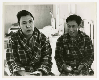 Two patients of the Hospital sitting on the same edge of a bed. Both patients are looking at opposite sides of the camera, out of the field of image. Both are dressed in pyjamas and plaid patterned housecoats. Each is holding a soapstone carving in their hands. Three more beds are visible, though out of focus, in the background with unidentified medical equipment. Two windows are visible.