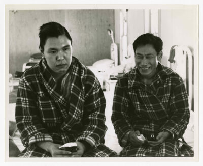 Two patients of the Hospital sitting on the same edge of a bed. Both patients are looking at opposite sides of the camera, out of the field of image. The man on the left has a blank look on his face, and the man on the right is smiling. Both are dressed in pyjamas and plaid patterned housecoats. Each is holding a soapstone carving in their hands. Three more beds are visible, though out of focus, in the background with unidentified medical equipment. Two windows are visible.