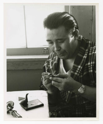 A man working on a very small piece of soapstone. He is wearing pyjamas, a plaid patterned housecoat and a wristwatch. On the table in front of him is a carved goose on a stand with what may be eggs. What may be a hand cranked drill and piece of white stone are also on the table. A closed window is visible directly behind the subject.
