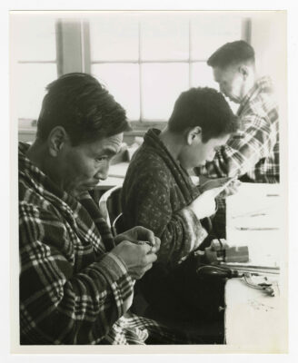Three men carving. The man on the left appears to be polishing an ivory ring while he eyes the camera out of the corner of his eye. Another man is wearing pyjamas and a plaid pattered housecoat at the back of the image. A teenager is in between the two men, and is working a small piece of soapstone with a round file. He is wearing a ring and polka-dotted patterned housecoat over his pyjamas. The table in the foreground has a variety of tools used for soapstone carving on them as well as a table-mounted vice.