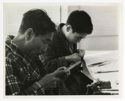 A man and a teenager carving. The man on the left appears to be filing an ivory ring. He is wearing pyjamas and a plaid pattered housecoat. The teenager to the right is working a small unknown piece. He is wearing a circle patterned housecoat over his pyjamas. The table that they are working at has a variety of tools on it as well as a table-mounted vice.