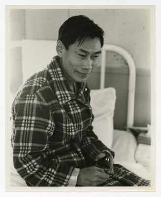 A man smiling and looking down towards the floor. He is sitting on a hospital bed holding a carved walrus. The walrus is a dark colour with white tusks and about the size of an adult fist. The man is dressed in pyjamas with a plaid patterned housecoat. The head of the bed is visible behind him and is up against a wall.
