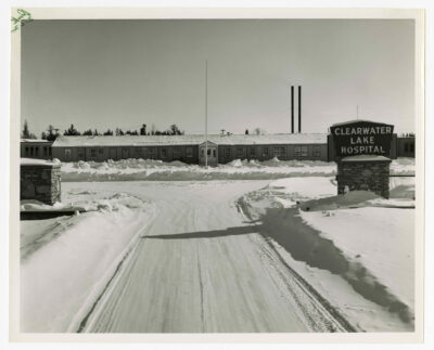 The entrance to Clearwater Lake Hospital. The front gate walls, sign, flagpole and front of the building are visible. There is snow piled up on the ground and the area for cars is packed with snow. There is no flag on the flagpole in front of the building. The building itself is one storey with many windows and stretches from one side of the photograph to the other. Two smokestacks are visible from behind the building in the PL half of the image. Trees are visible in the very back of the image.